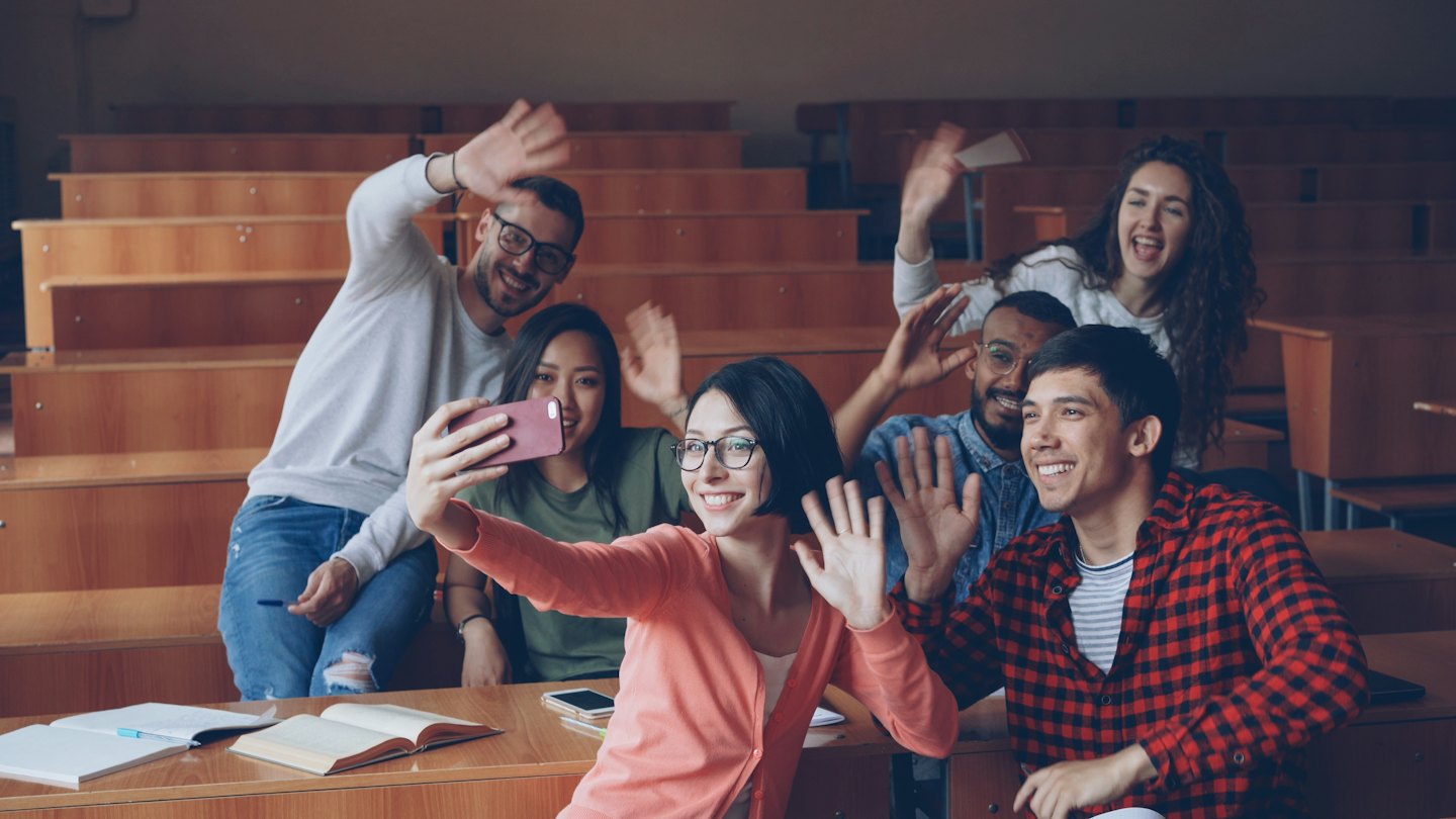 Studenti salutano verso la telecamera in un'aula universitaria.
